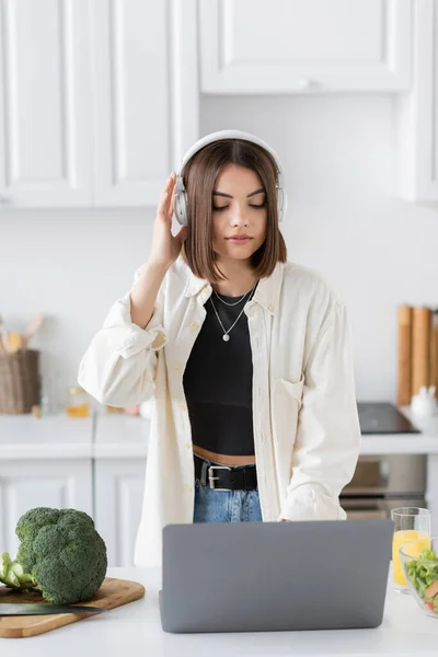 Brunette woman in headphones using laptop near vegetables and fresh salad in kitchen — Stock Photo