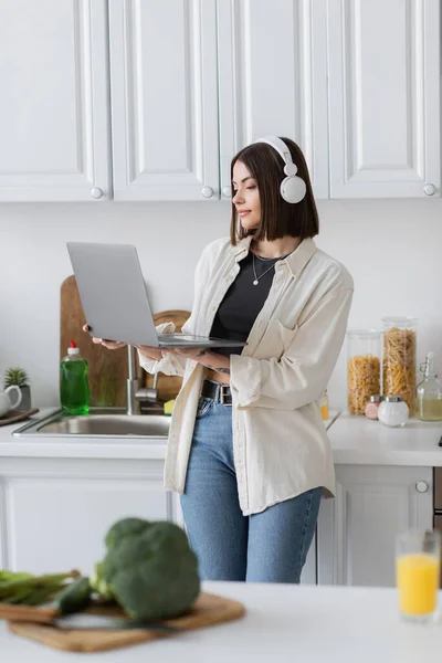 Jovem mulher em fones de ouvido usando laptop perto de legumes desfocados e suco de laranja na cozinha — Fotografia de Stock