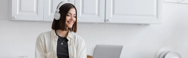 Femme brune souriante dans les écouteurs en utilisant un ordinateur portable dans la cuisine à la maison, bannière — Photo de stock