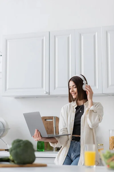 Mulher positiva em fones de ouvido usando laptop perto de brócolis borrado e suco de laranja na cozinha — Fotografia de Stock