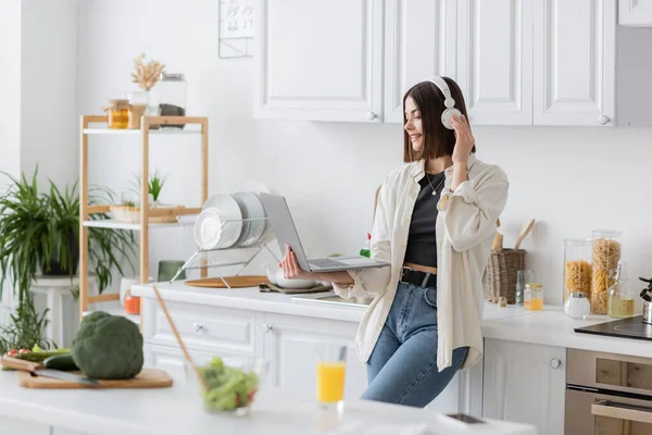 Mujer sonriente en auriculares inalámbricos con computadora portátil cerca de ensalada borrosa y jugo de naranja en la cocina - foto de stock