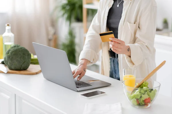 Vue recadrée d'une femme tatouée tenant une carte de crédit et utilisant un ordinateur portable près d'une salade floue dans la cuisine — Photo de stock
