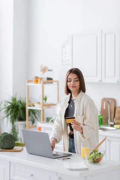 Brunette woman holding credit card and using laptop while ordering food in kitchen — Stock Photo