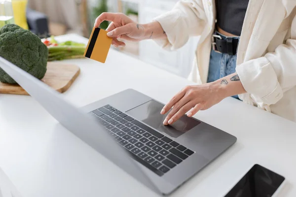 Vue recadrée d'une femme tatouée tenant une carte de crédit et utilisant un ordinateur portable près de légumes mûrs dans la cuisine — Photo de stock