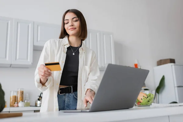 Mujer morena sosteniendo tarjeta de crédito cerca de la computadora portátil y ensalada fresca en la cocina - foto de stock