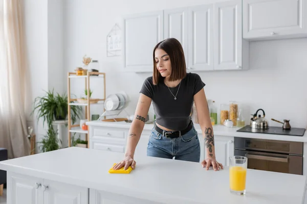 Tattooed brunette woman cleaning worktop with rag in kitchen — Stock Photo