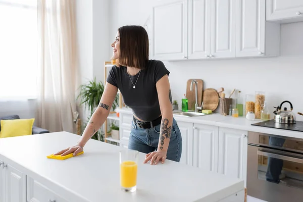 Vista laterale della donna tatuata sorridente che pulisce il piano di lavoro con lo straccio vicino al succo d'arancia in cucina — Foto stock
