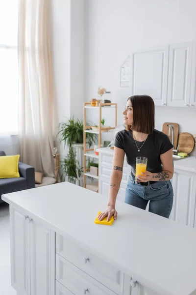 Side view of tattooed woman smiling and holding orange juice while cleaning worktop in kitchen — Stock Photo