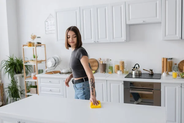 Joven mujer tatuada limpiando encimera con trapo en la cocina en casa - foto de stock