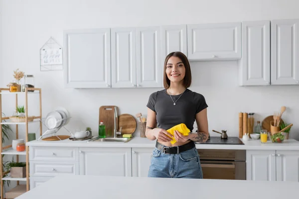 Sorglos tätowierte Frau hält Lappen in der Hand und blickt in die Küche in die Kamera — Stockfoto