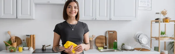 Mujer morena sonriente sosteniendo trapo mientras limpia la cocina en casa, pancarta - foto de stock