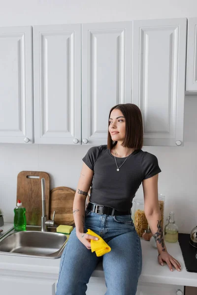 Усміхнена брюнетка жінка тримає ганчірку, сидячи на робочому столі на кухні — стокове фото