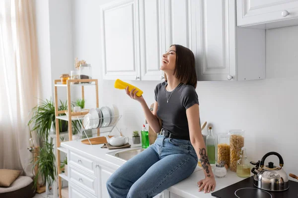 Überglücklich tätowierte Frau hält Lappen in der Hand, während sie auf Arbeitsplatte in der Küche sitzt — Stockfoto