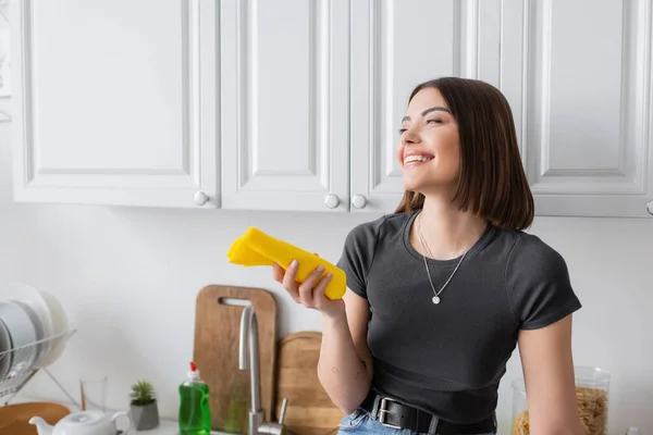 Mujer morena sonriente sosteniendo trapo mientras está de pie en la cocina en casa - foto de stock