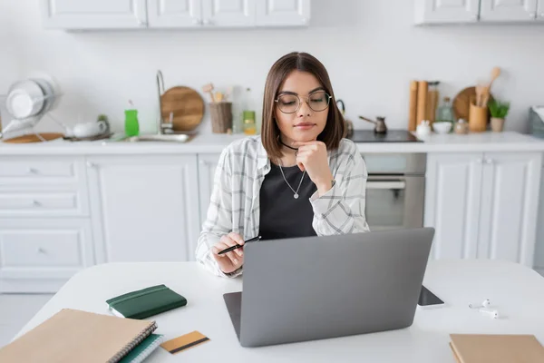 Brunette freelancer looking at laptop near notebooks and credit card in kitchen — Stock Photo