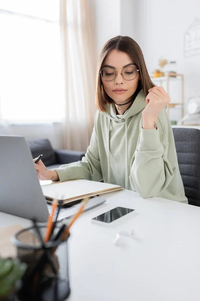 Freelancer in eyeglasses looking at smartphone near notebook and laptop at home — Stock Photo