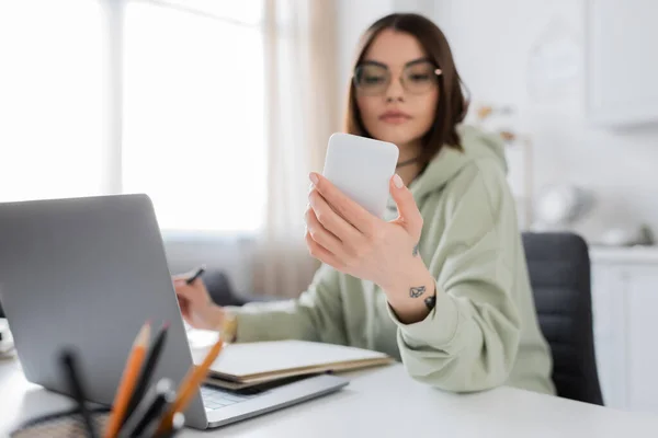 Blurred freelancer holding cellphone near laptop and notebook at home — Stock Photo