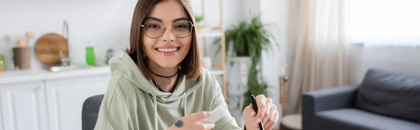 Усміхнена жінка в окулярах тримає смартфон і ручку вдома, банер — стокове фото