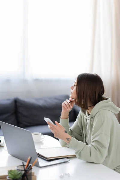 Vista lateral del freelancer pensativo sosteniendo el teléfono celular cerca de la computadora portátil y portátil en casa - foto de stock