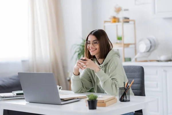 Freelancer sonriente en gafas con bolígrafo cerca del portátil y cuadernos en casa - foto de stock