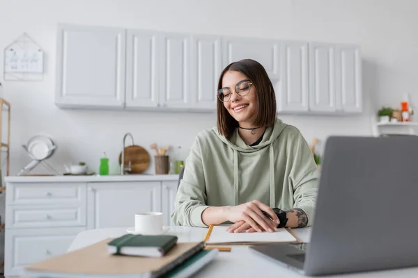 Freelancer positivo em óculos olhando para xícara de café perto de laptop e notebooks na mesa em casa — Fotografia de Stock