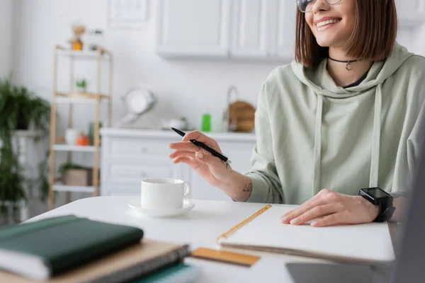 Cropped view of positive freelancer holding pen near coffee and notebooks at home — Stock Photo