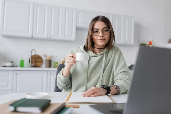 Freelancer tatuado em óculos segurando café perto de laptop desfocado e notebooks em casa — Fotografia de Stock