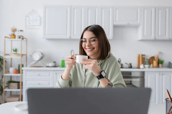 Freelance positif dans les lunettes de vue tenant café près d'un ordinateur portable flou à la maison — Photo de stock