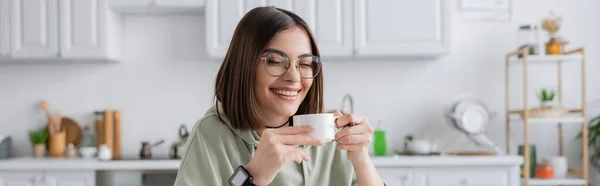 Весела молода жінка в окулярах тримає каву в розмитій кухні, банер — стокове фото