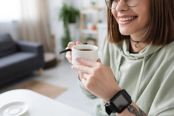 Vista cortada de mulher tatuada sorridente com relógio inteligente segurando café e caneta em casa — Fotografia de Stock