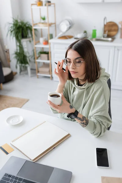 Tätowierte Frau mit Brille hält Kaffeetasse in der Nähe von Geräten und Kreditkarte zu Hause auf dem Tisch — Stockfoto