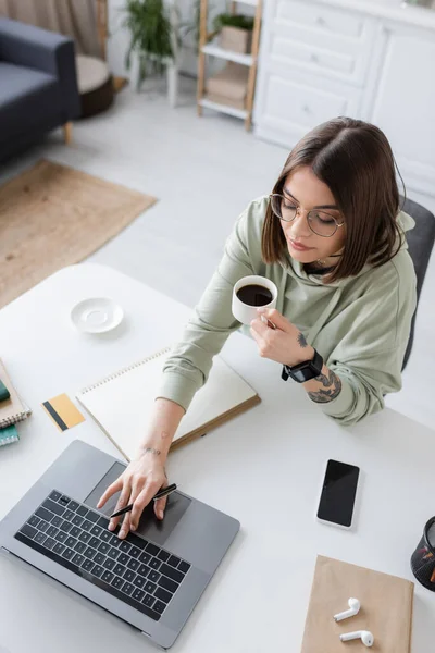 Overhead view of tattooed freelancer holding coffee and using laptop near notebooks and earphones at home — Stock Photo