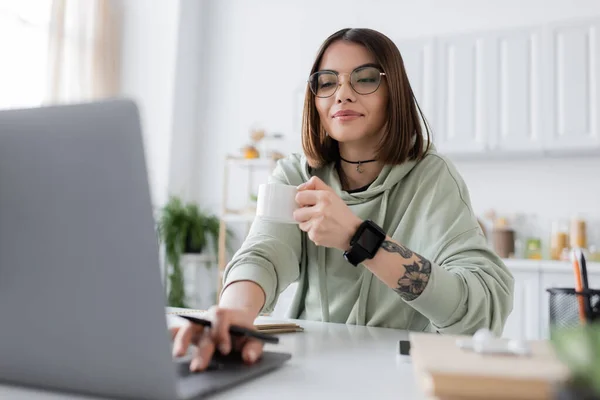 Freelancer tatuado em óculos segurando xícara de café e usando laptop desfocado em casa — Fotografia de Stock