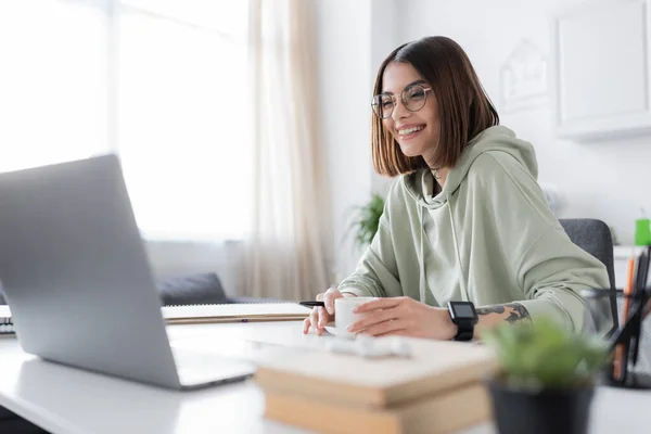 Cheerful freelancer in eyeglasses looking at laptop near coffee and books at home — Stock Photo