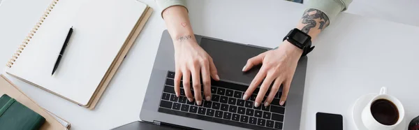 Вид сверху на татуированного фрилансера, использующего ноутбук рядом с тетрадями и чашкой кофе на столе, баннер — стоковое фото