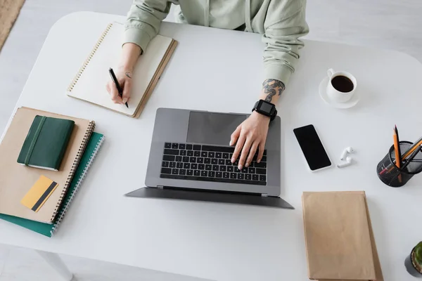 Vista ritagliata di freelance utilizzando laptop e scrittura su notebook vicino a caffè e auricolari sul tavolo a casa — Foto stock