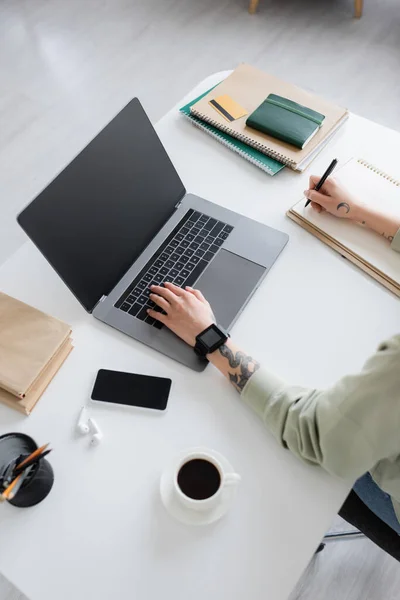 Top view of tattooed freelancer writing on notebook and using laptop near coffee and books on table — Stock Photo