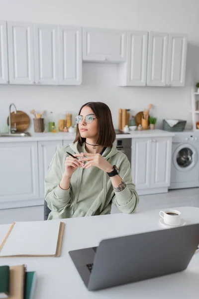Молодой фрилансер в очках держит ручку рядом с ноутбуком и ноутбуком на столе дома — стоковое фото