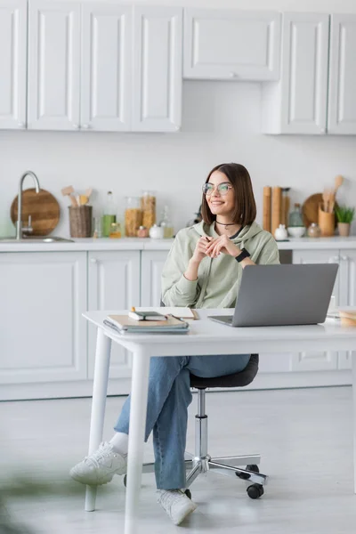 Femme souriante assise près d'un ordinateur portable et des cahiers sur la table dans la cuisine — Photo de stock