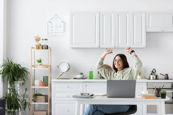 Aufgeregte Freiberuflerin zeigt Ja-Geste neben Laptop und Notizbuch auf Tisch in Küche — Stockfoto