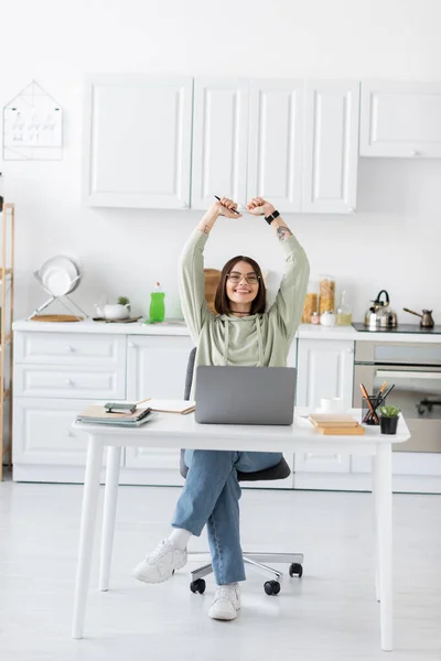 Indépendante excitée dans les lunettes montrant un geste oui près d'un ordinateur portable et des ordinateurs portables dans la cuisine — Photo de stock