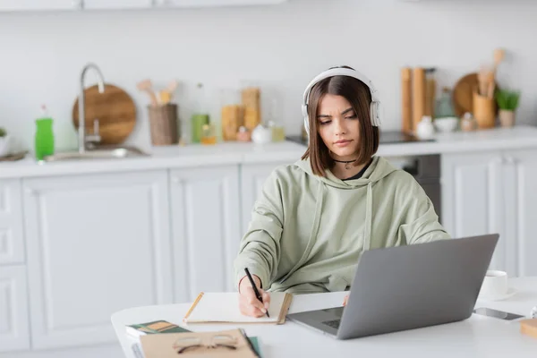 Jeune pigiste dans un casque d'écriture sur ordinateur portable près de gadgets et café sur la table à la maison — Photo de stock
