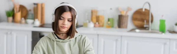 Brunette woman in headphones looking away in blurred kitchen, banner — Stock Photo