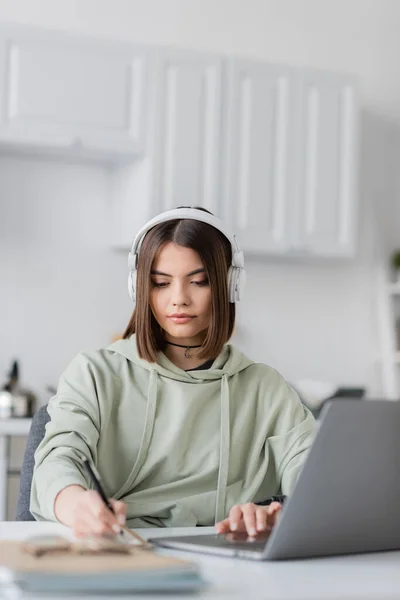 Brunette freelancer in headphones using laptop and blurred notebook at home — Stock Photo