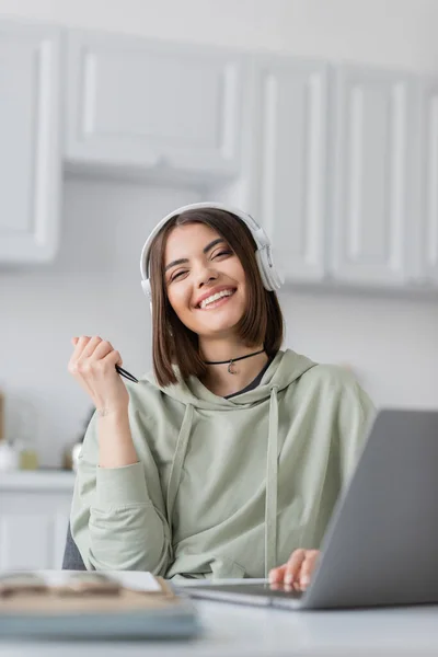 Carefree freelancer in headphones looking at camera near laptop and notebooks at home — Stock Photo