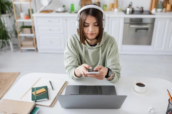 Freelancer morena em fones de ouvido usando smartphone perto de laptop e café em casa — Fotografia de Stock