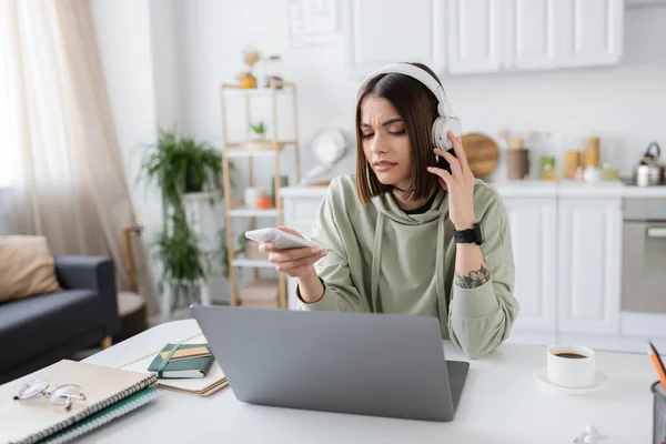Displeased freelancer in headphones holding cellphone near laptop and coffee at home — Stock Photo