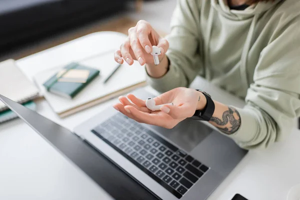 Cropped view of tattooed freelancer holding earphones near laptop with blank screen on table at home — Stock Photo