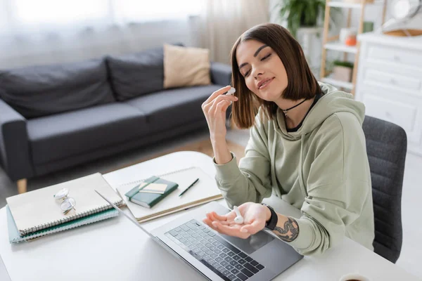 Smiling tattooed freelancer holding earphones near laptop and notebooks at home — Stock Photo