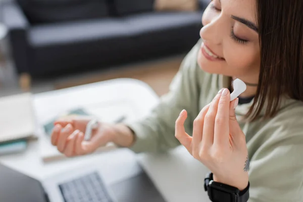 Cheerful freelancer holding earphone near blurred laptop at home — Stock Photo